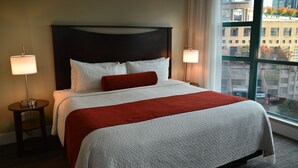 A hotel room with a large bed, two bedside lamps, a window with a city view, and a dark headboard. - Rosedale Condominiums (Vancouver)