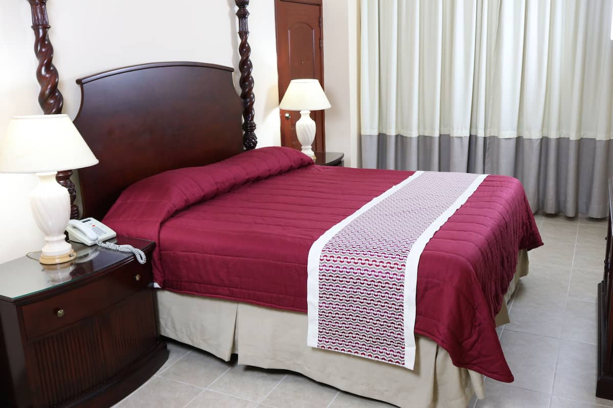 Una cama bien tendida con una colcha roja y un borde decorativo blanco, una mesita de noche de madera con una lámpara y un teléfono, y una puerta con un cortinaje de fondo.
