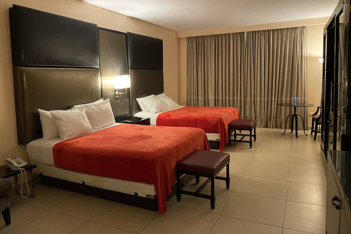 Habitación de hotel con dos camas, una mesita de noche con lámpara, una pequeña mesa con silla y un ventanal con cortinas.