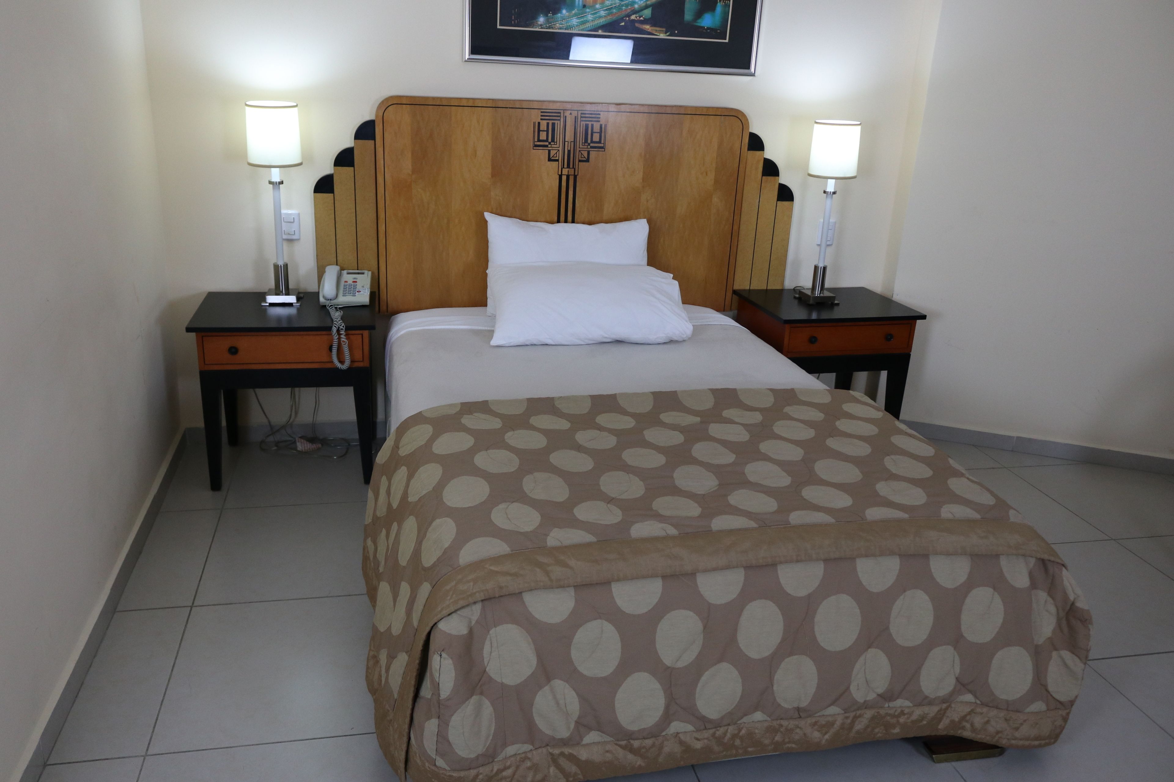 A hotel room with a bed, two bedside tables, a nightstand, a lamp, a phone, and a framed picture on the wall.
