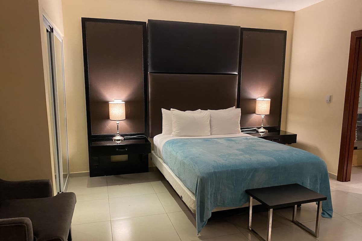 Una habitación de hotel con una cama grande, dos mesitas de noche con lámparas, un banco y una silla.
