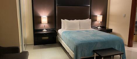 Quarto de hotel com uma cama grande, duas mesas de cabeceira com luminárias, um banco e uma cadeira.