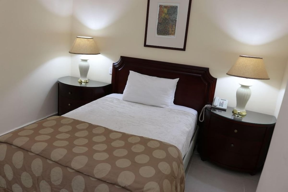 Una habitación de hotel con cama, dos mesitas de noche, un atril con teléfono y un cuadro en la pared.