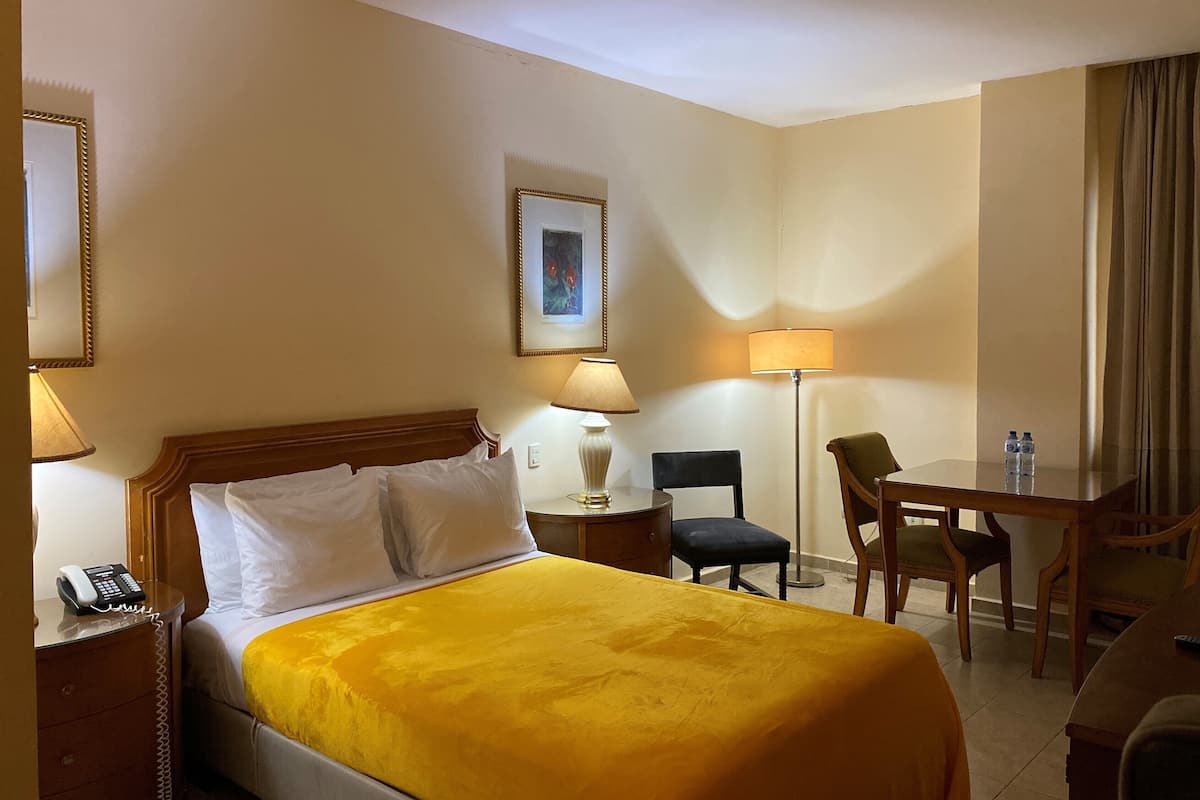 Una habitación de hotel con cama, mesita de noche con teléfono, escritorio con dos sillas, lámpara y un cuadro en la pared.