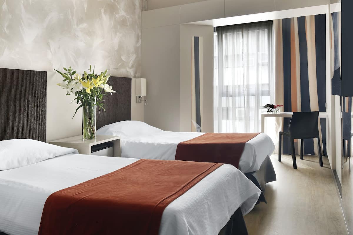 A hotel room with two beds, a desk, a chair, and a vase with flowers.