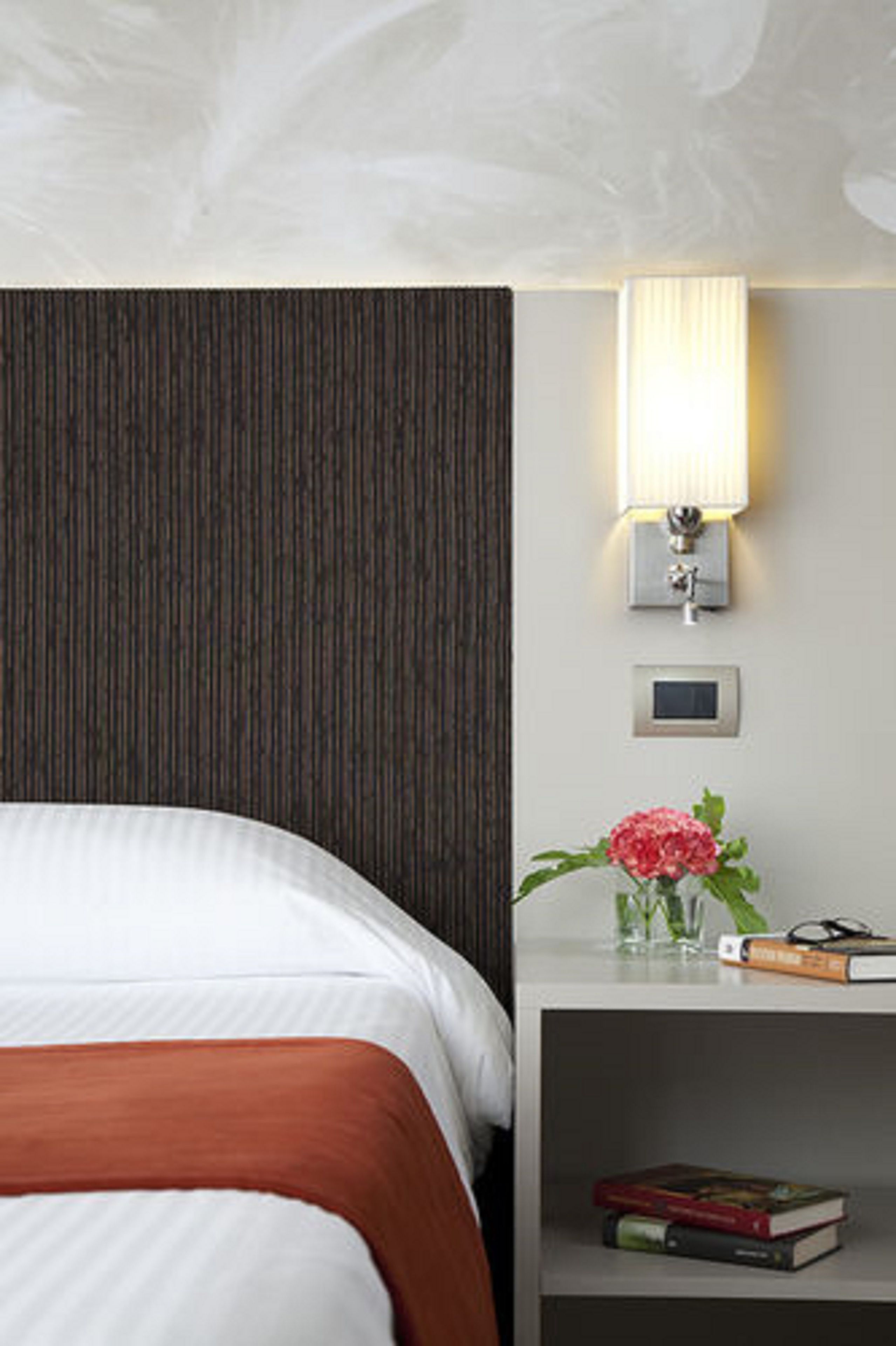 A hotel room with a bed, a nightstand with a lamp, a small vase with flowers, and a book.