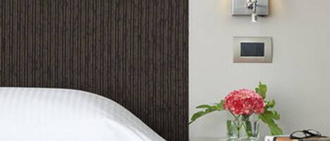 A hotel room with a bed, a nightstand with a lamp, a small vase with flowers, and a book.