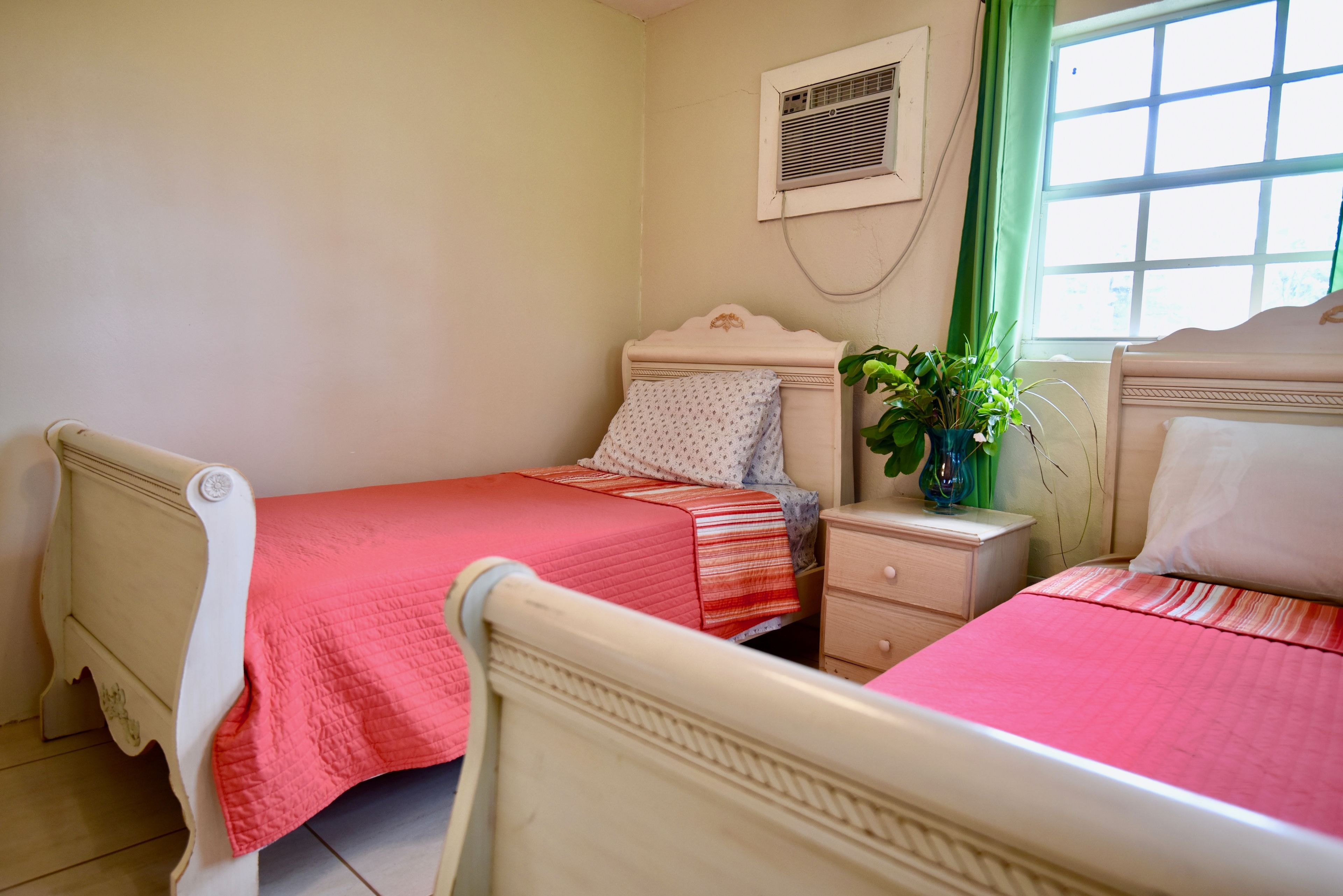 Une chambre avec deux lits, une table de chevet, une commode, une plante en pot et une fenêtre avec des rideaux.