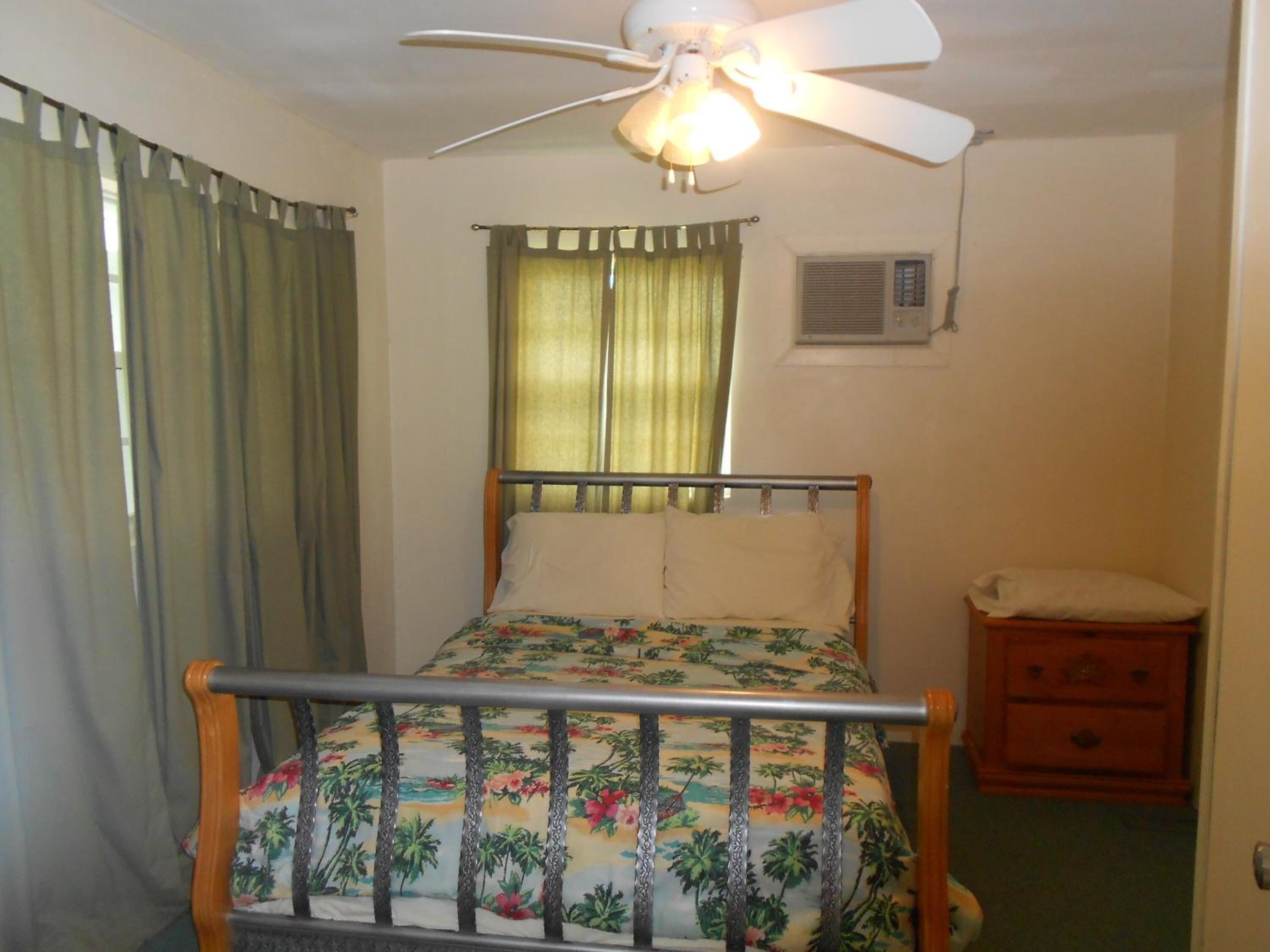 Une chambre à coucher avec un lit, une table de chevet en bois, un ventilateur de plafond et une fenêtre avec des rideaux.