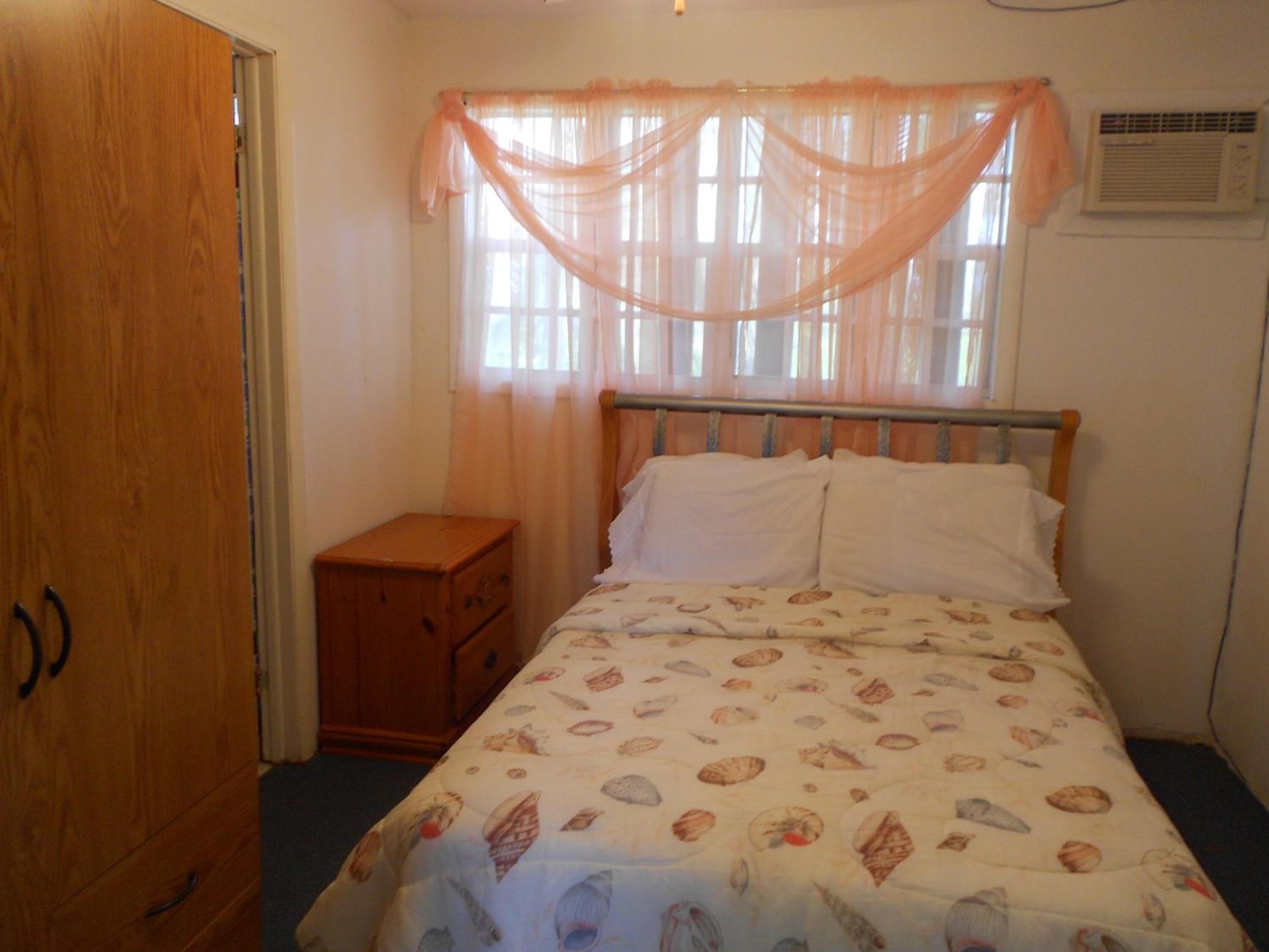 Une chambre à coucher comprenant un lit, une armoire en bois, une table de chevet et une fenêtre avec des rideaux légers.