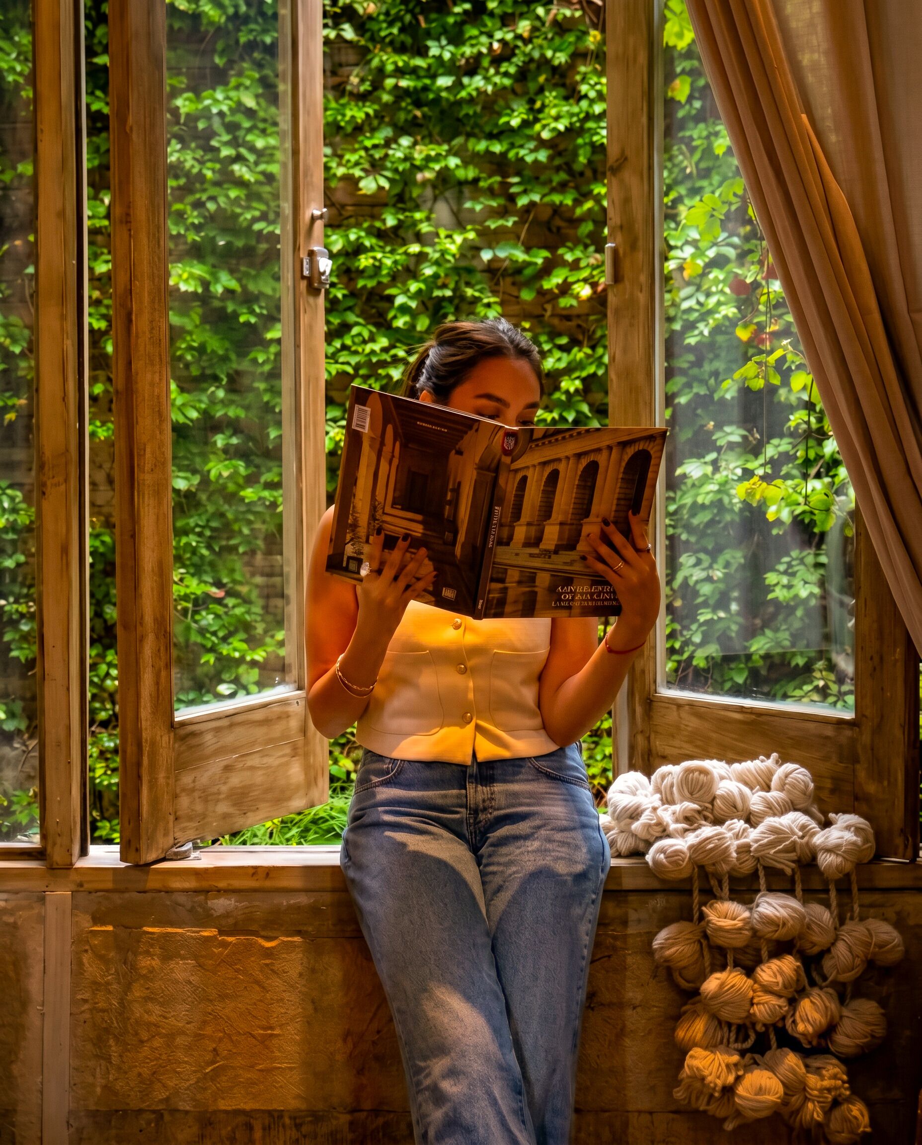 Una persona leyendo un libro junto a una ventana con vistas a zonas verdes.