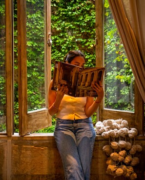 Une personne est assise près d’une fenêtre, un livre à la main, et la vue donne sur un paysage verdoyant.