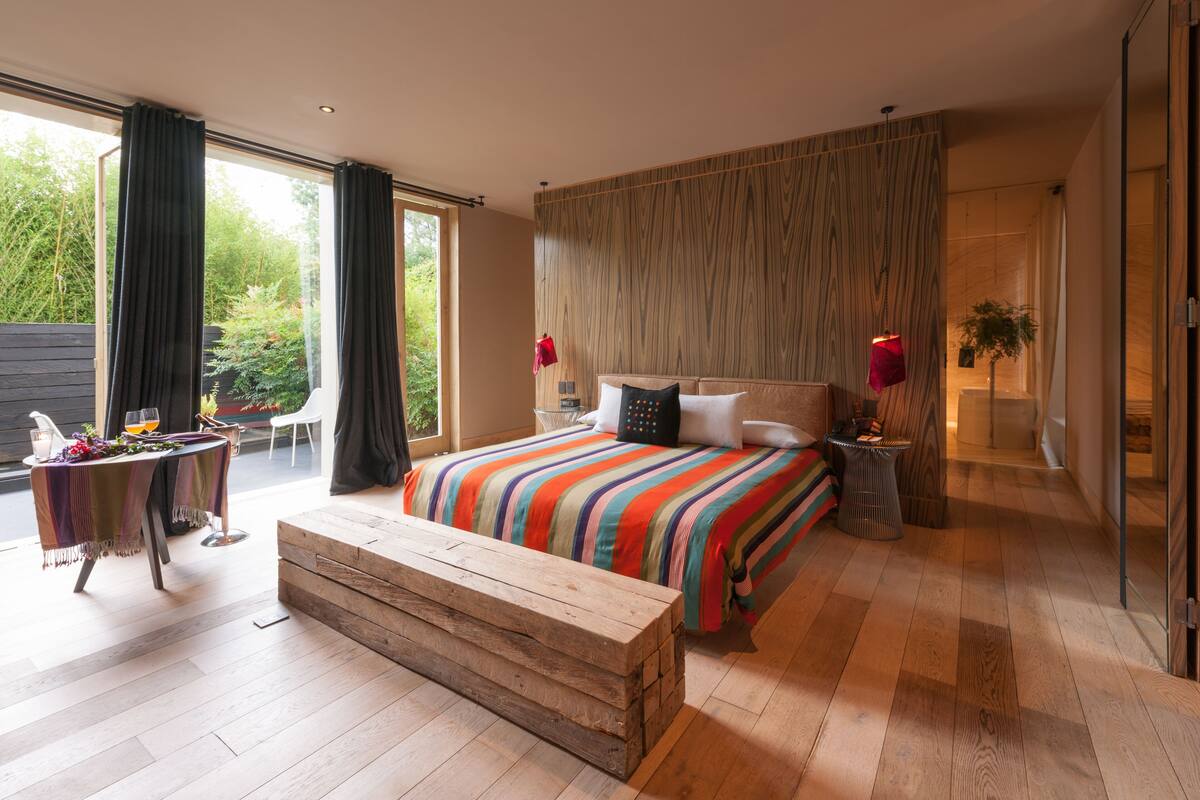 Une chambre avec un grand lit, un plancher en bois, un banc, une table avec des boissons et une vue sur la verdure à l’extérieur.
