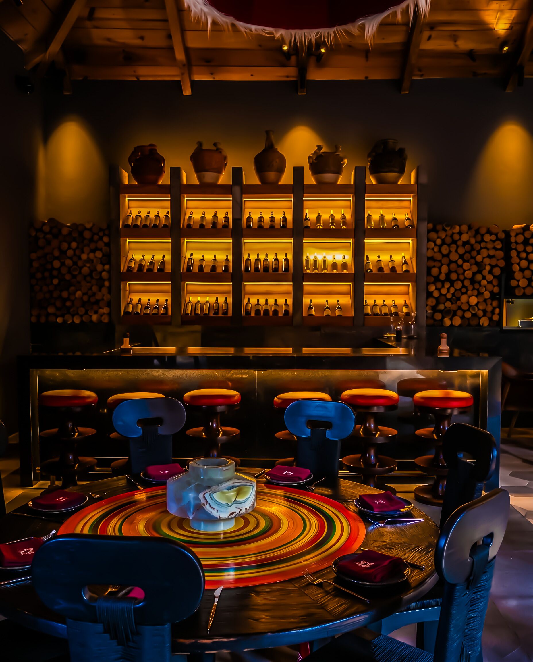 Una zona de bar con estanterías llenas de botellas, una mesa redonda con platos de colores y taburetes.