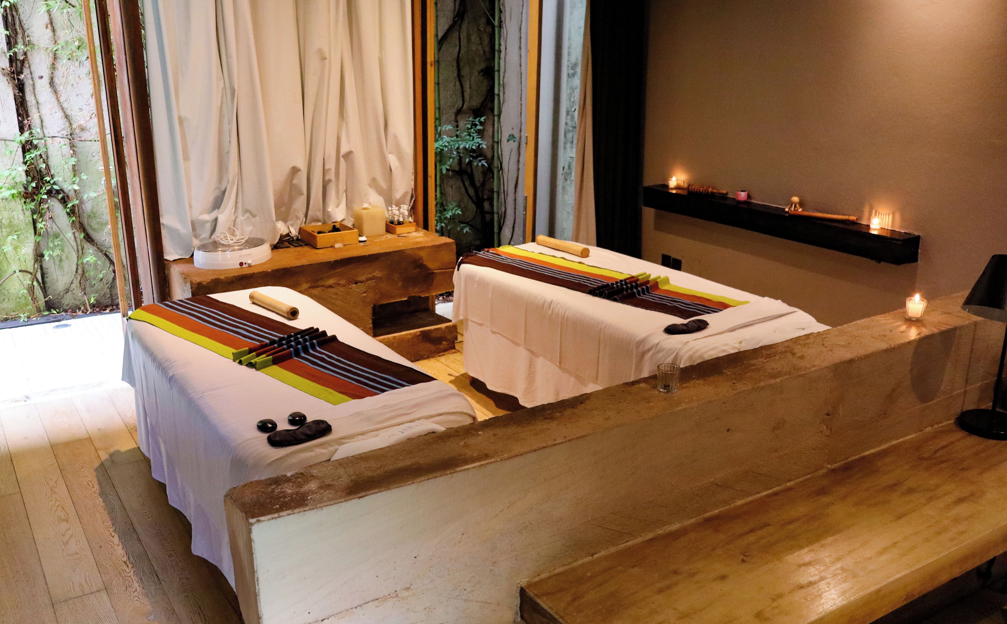 Een kamer met twee massagetafels, een houten bank, een plank met kaarsen, en een raam met gordijnen.