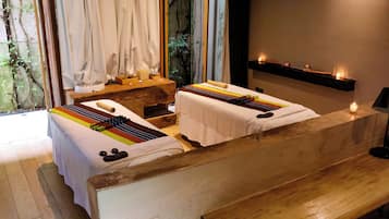 Een kamer met twee massagetafels, een houten bank, een plank met kaarsen, en een raam met gordijnen.