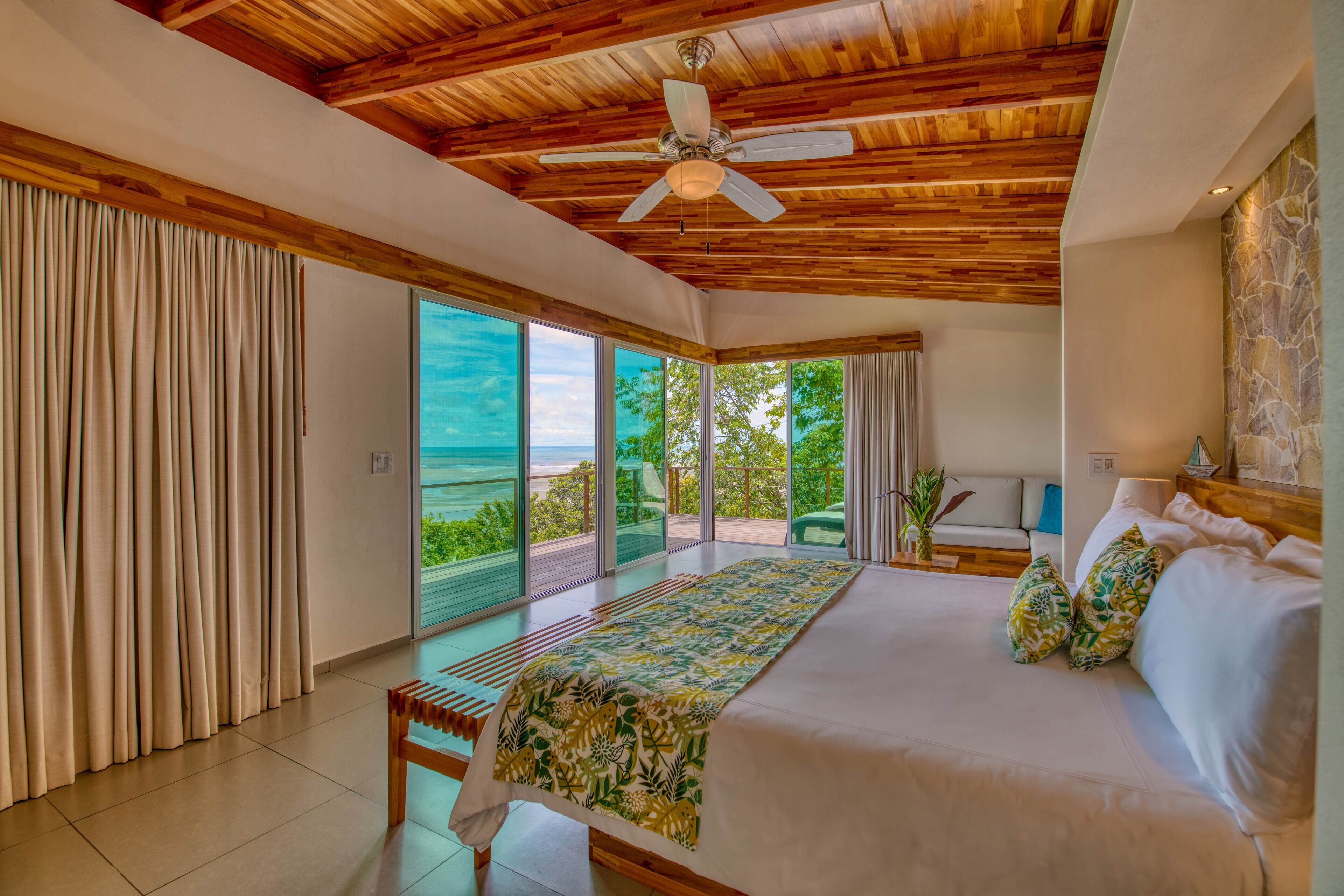 A spacious bedroom with a large bed, a ceiling fan, and a view of the ocean through sliding glass doors.