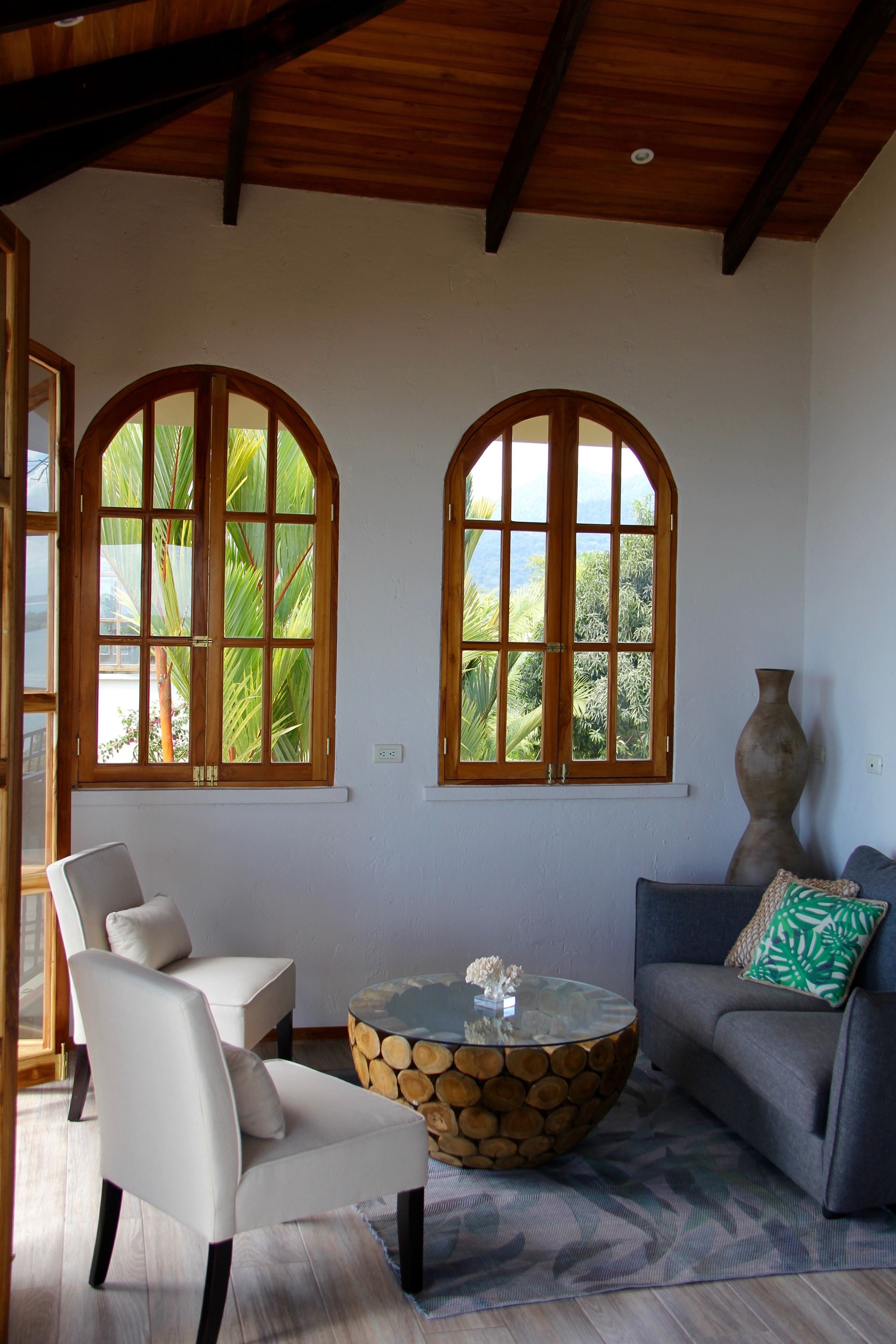 A room with two arched windows, a round coffee table with a log base, a white armchair, and a gray sofa with a green pillow.