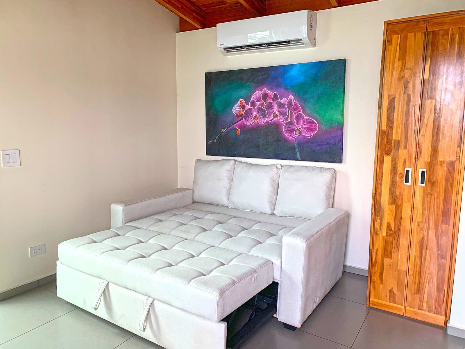 Una habitación con una cama, un equipo de aire acondicionado, un armario de madera y un cuadro de orquídeas.