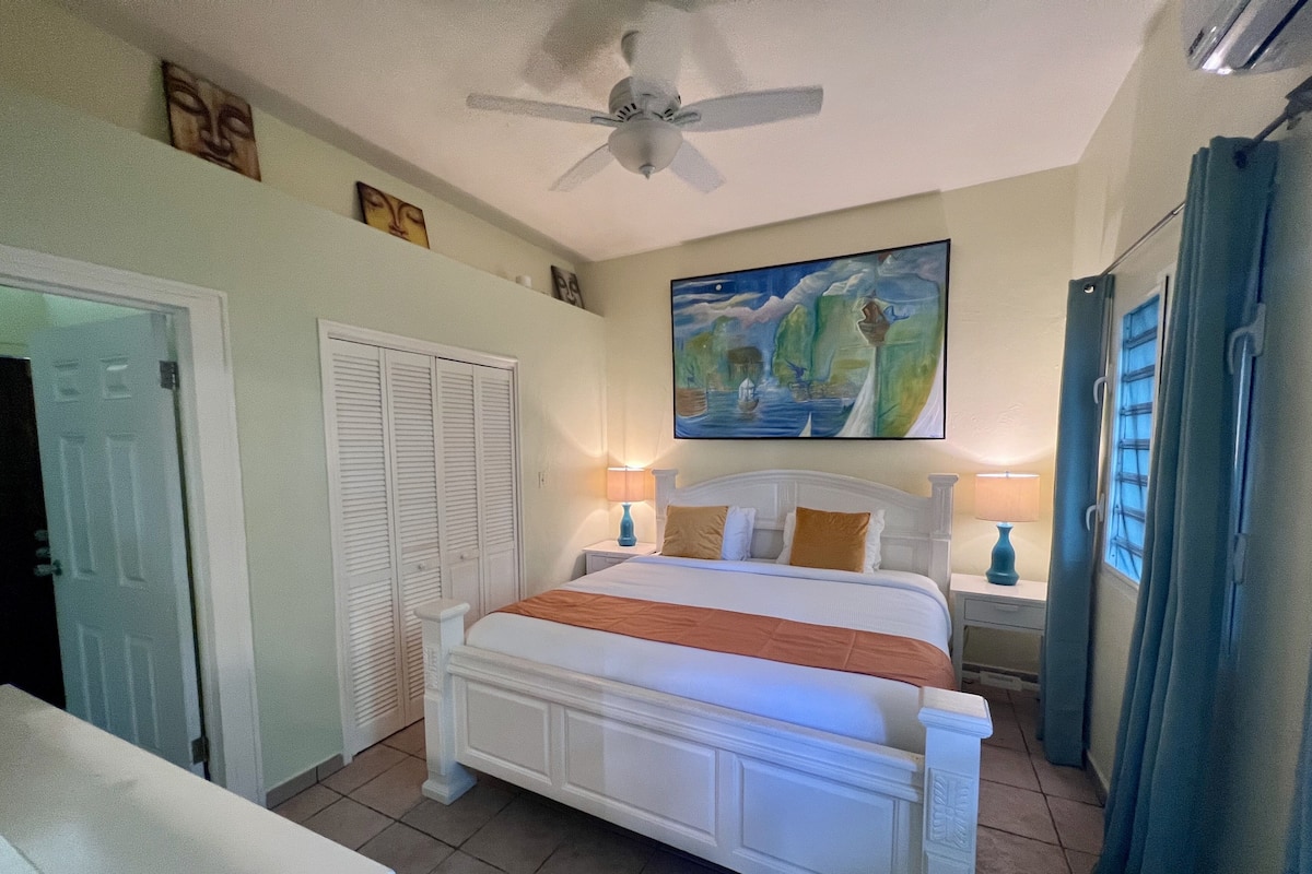 A bedroom with a bed, a ceiling fan, a painting, and a window with curtains.