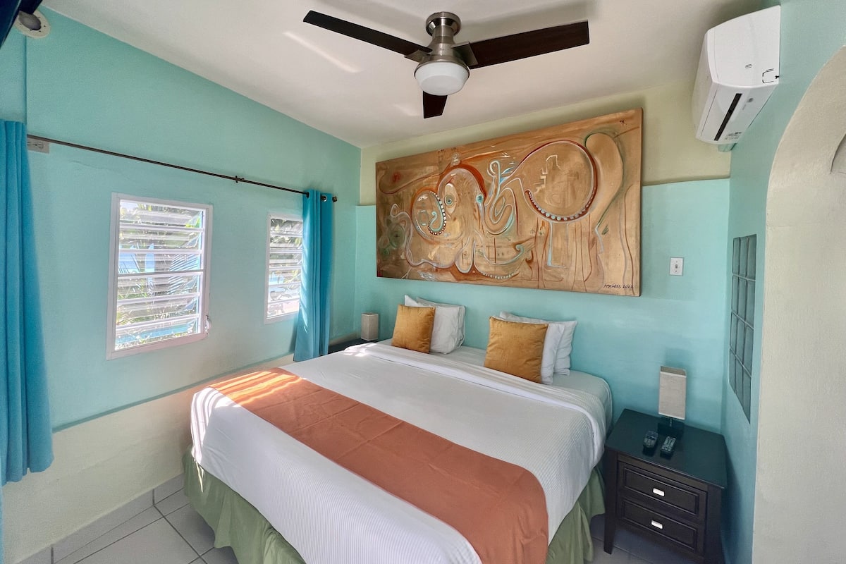 A bedroom with a bed, a ceiling fan, a wall-mounted air conditioner, and a window with curtains.