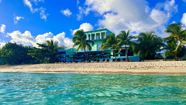 Un hôtel situé en bord de mer, doté d’une piscine, de palmiers et surplombant un ciel bleu azur.