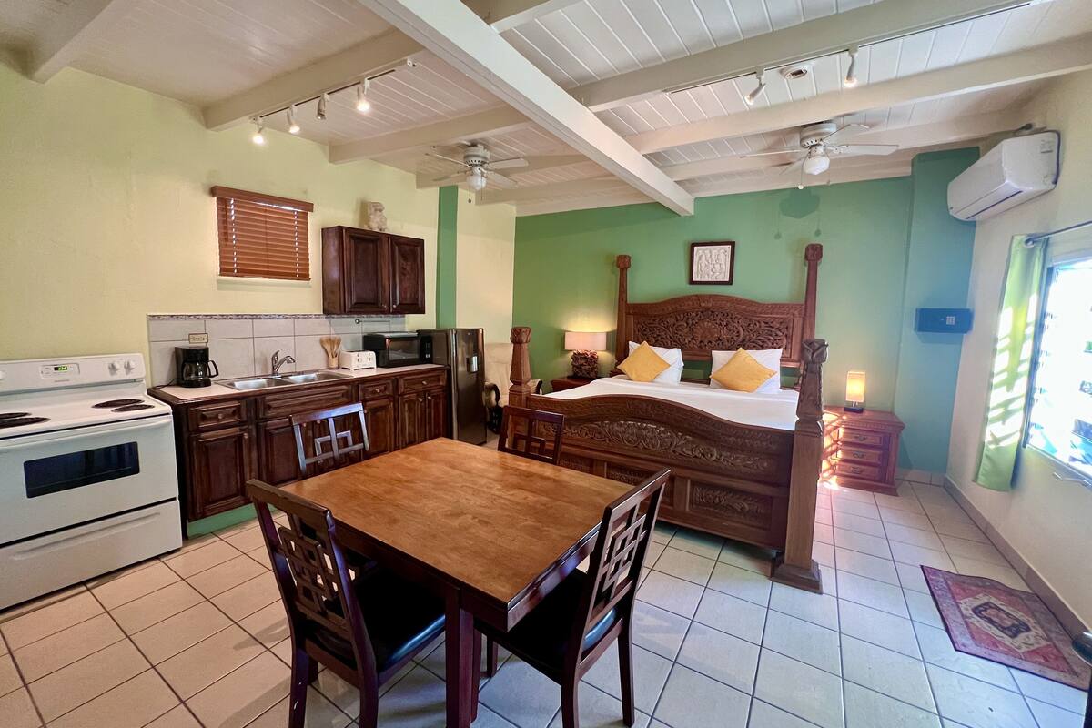 A kitchen with a stove, microwave, and refrigerator, a dining table with chairs, a bed with a wooden headboard, and a ceiling fan.