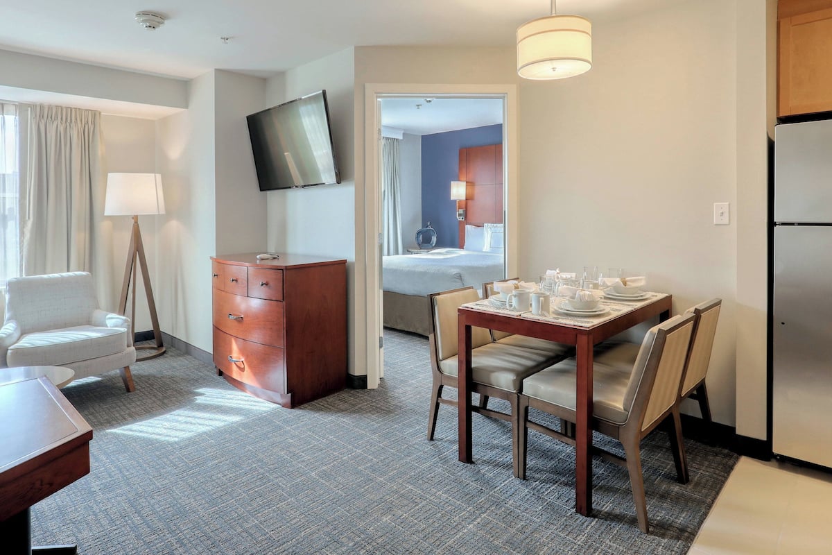 A hotel room with a dining area, a TV mounted on the wall, a wooden dresser, a bed with a blue headboard, and a refrigerator.
