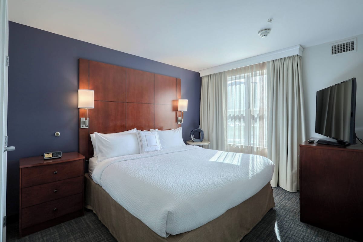 A hotel room with a large bed, a wooden headboard, two bedside lamps, a TV on a wooden cabinet, and a small table with a lamp.