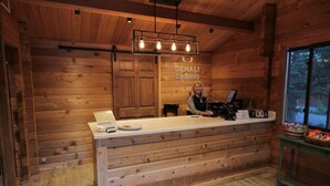 A wooden cabin interior with a reception desk, a sign reading "DENALI CABINS," and a window providing a view of the outdoors.