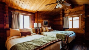 A cabin-style room with two beds, wooden walls, a ceiling fan, and large windows.