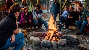 A group of people gathered around a campfire in an outdoor setting with wooden structures and trees in the background.