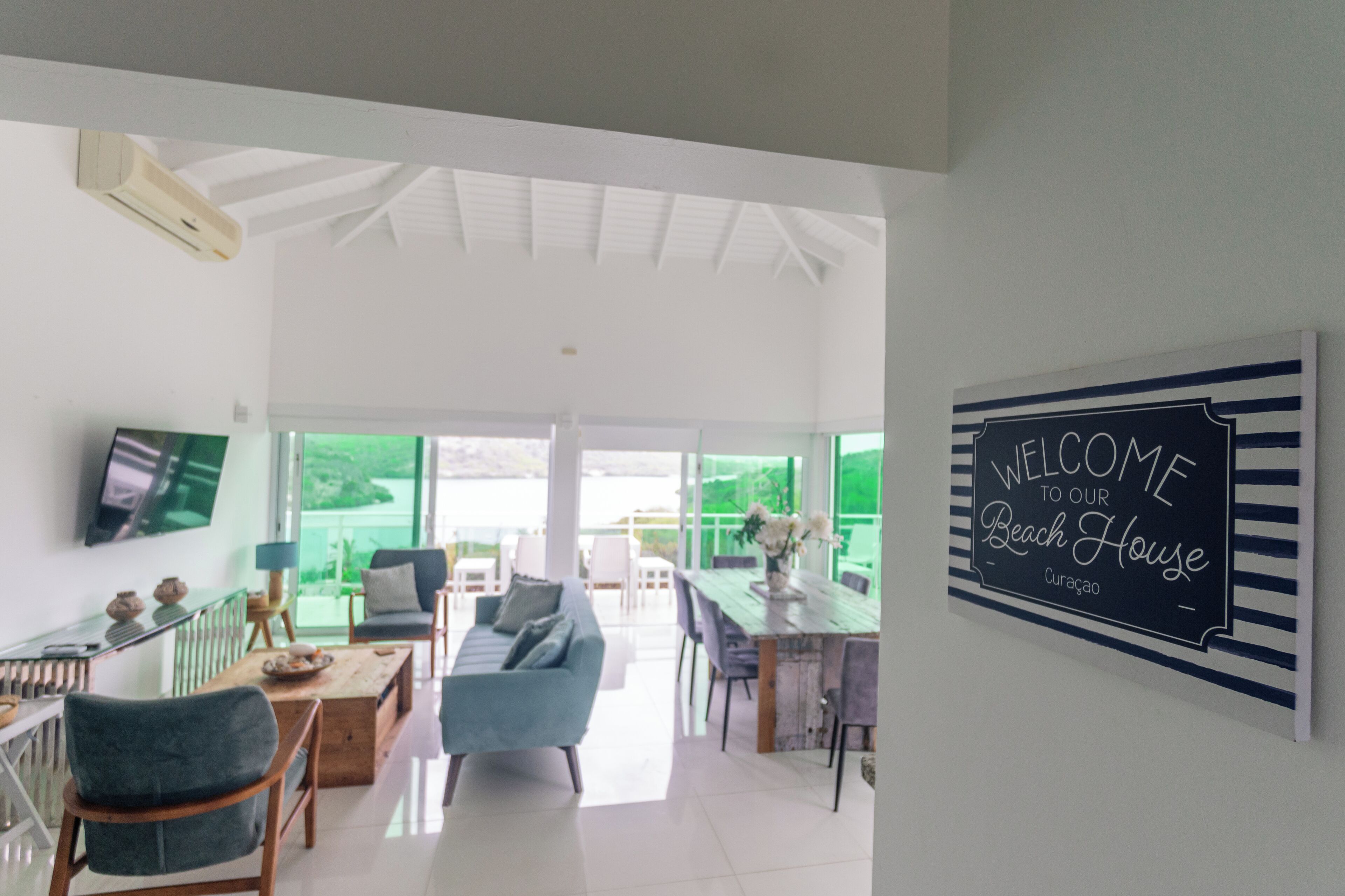 Un salón amplio con un diseño moderno, que incluye un sofá, una mesa de comedor y un televisor montado en la pared. Un cartel de bienvenida se muestra en la pared.