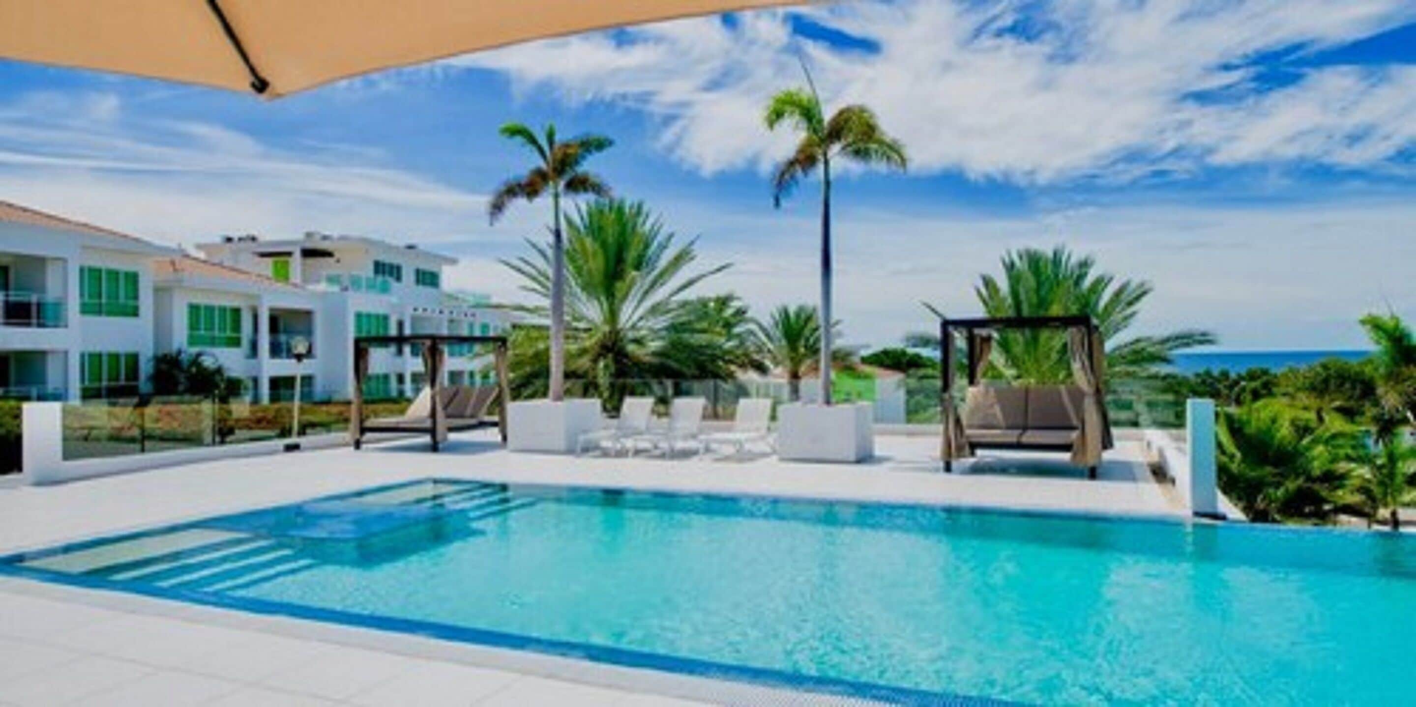 A swimming pool with lounge chairs and palm trees, surrounded by modern buildings with large windows.