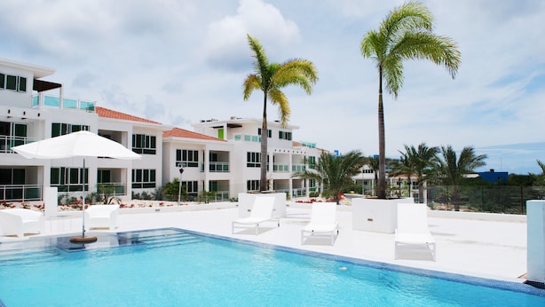 A swimming pool with white lounge chairs and umbrellas, surrounded by palm trees and modern buildings. - Piscadera Harbour Village (Willemstad)