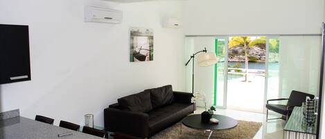 A modern living room with a dark brown sofa, a glass dining table, and a coffee table. There is a large window with a view of a natural landscape.
