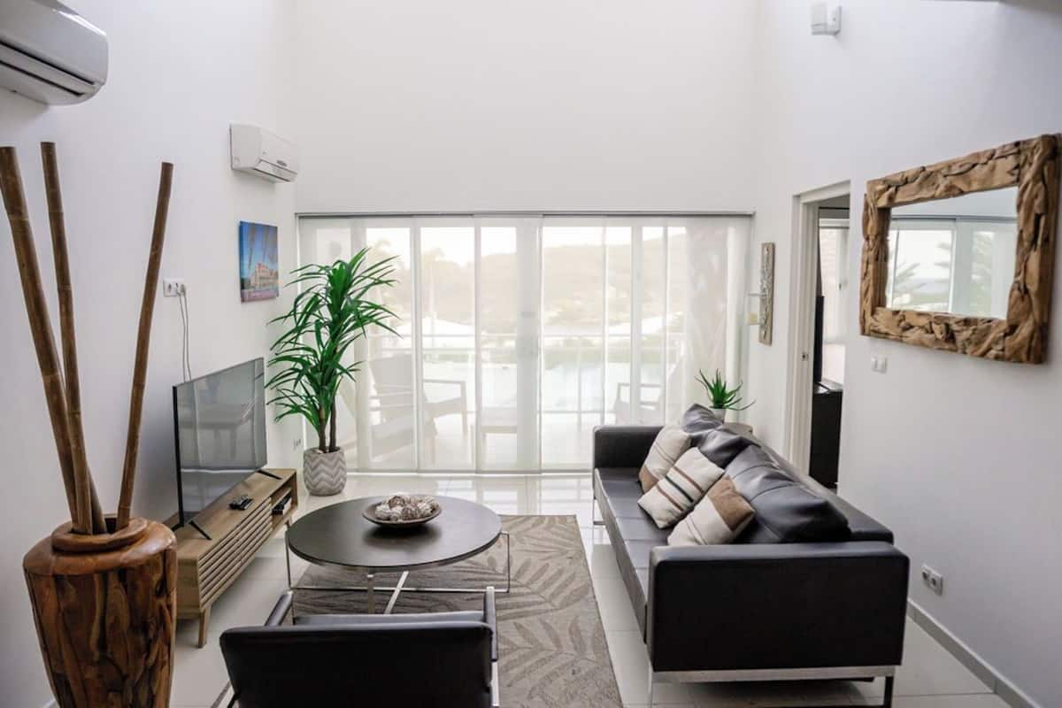 A modern living room with a sofa, a coffee table, and a TV. There are plants, a mirror, and a sliding glass door leading to a balcony.