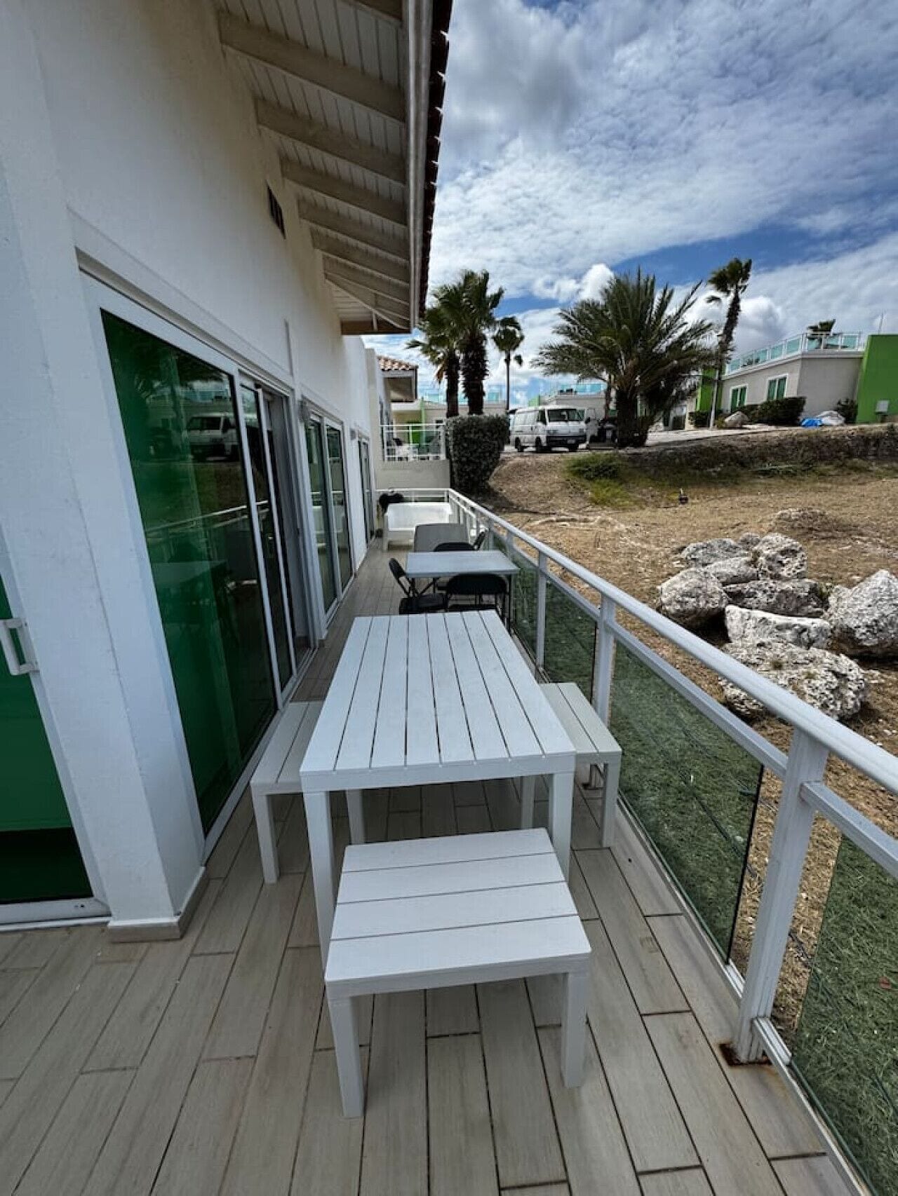 A covered outdoor area with a white table and bench, a glass door, and a view of a green building and palm trees.