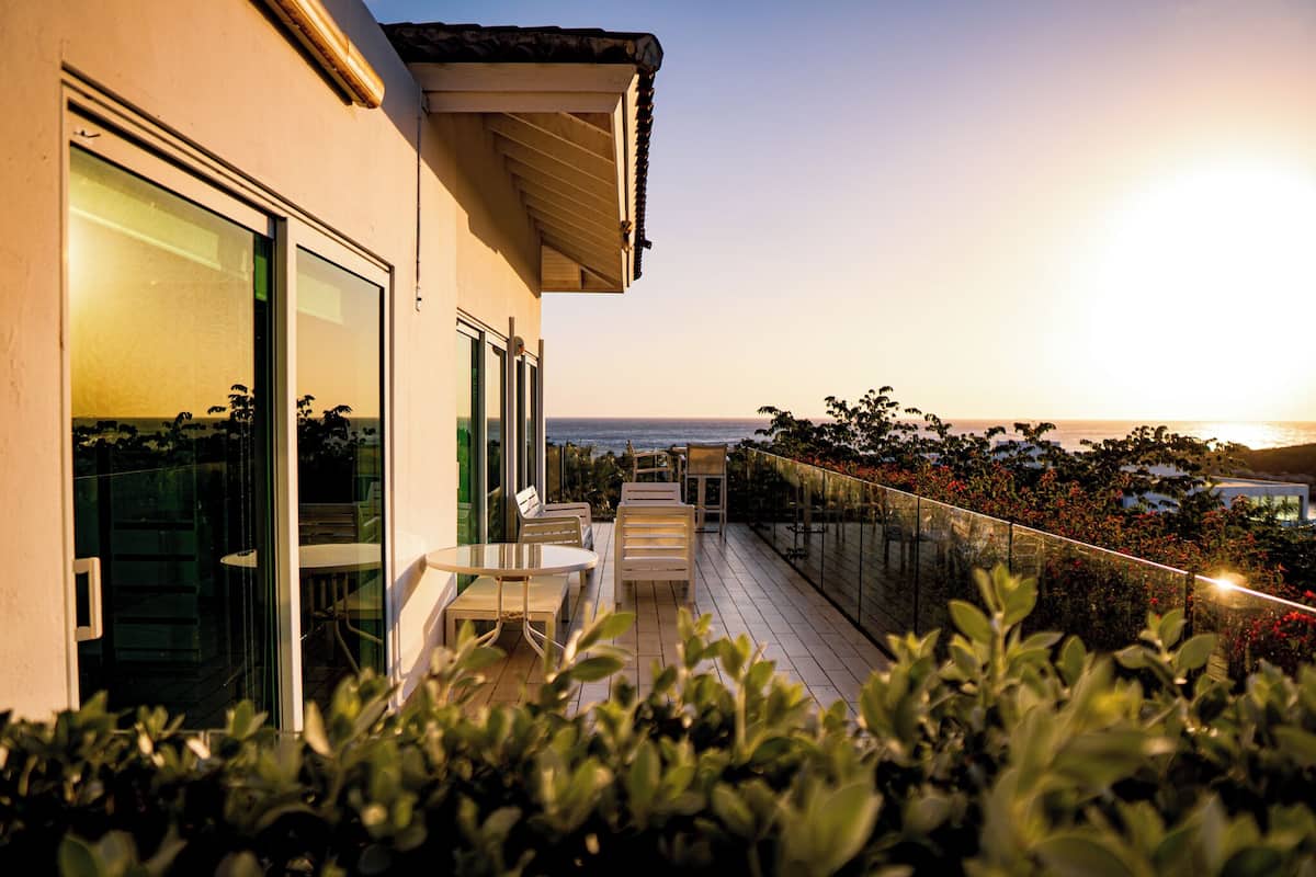 A balcony with outdoor seating, a glass door, and a view of the ocean at sunset.