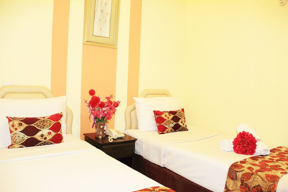 A hotel room with two beds, each with a white bedspread and red patterned pillows. There is a nightstand with a telephone and a vase of flowers between the beds.