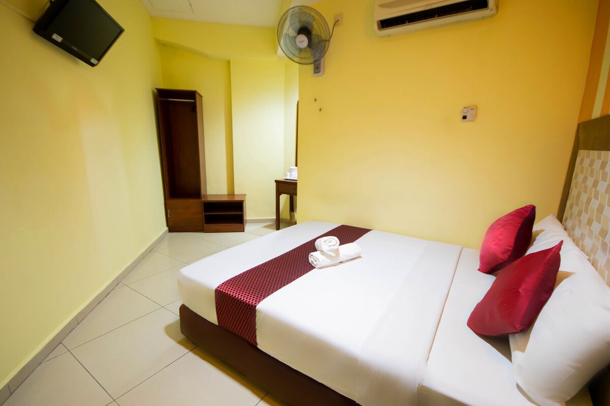 A hotel room with a bed, a bedside table, a fan, and an air conditioning unit.