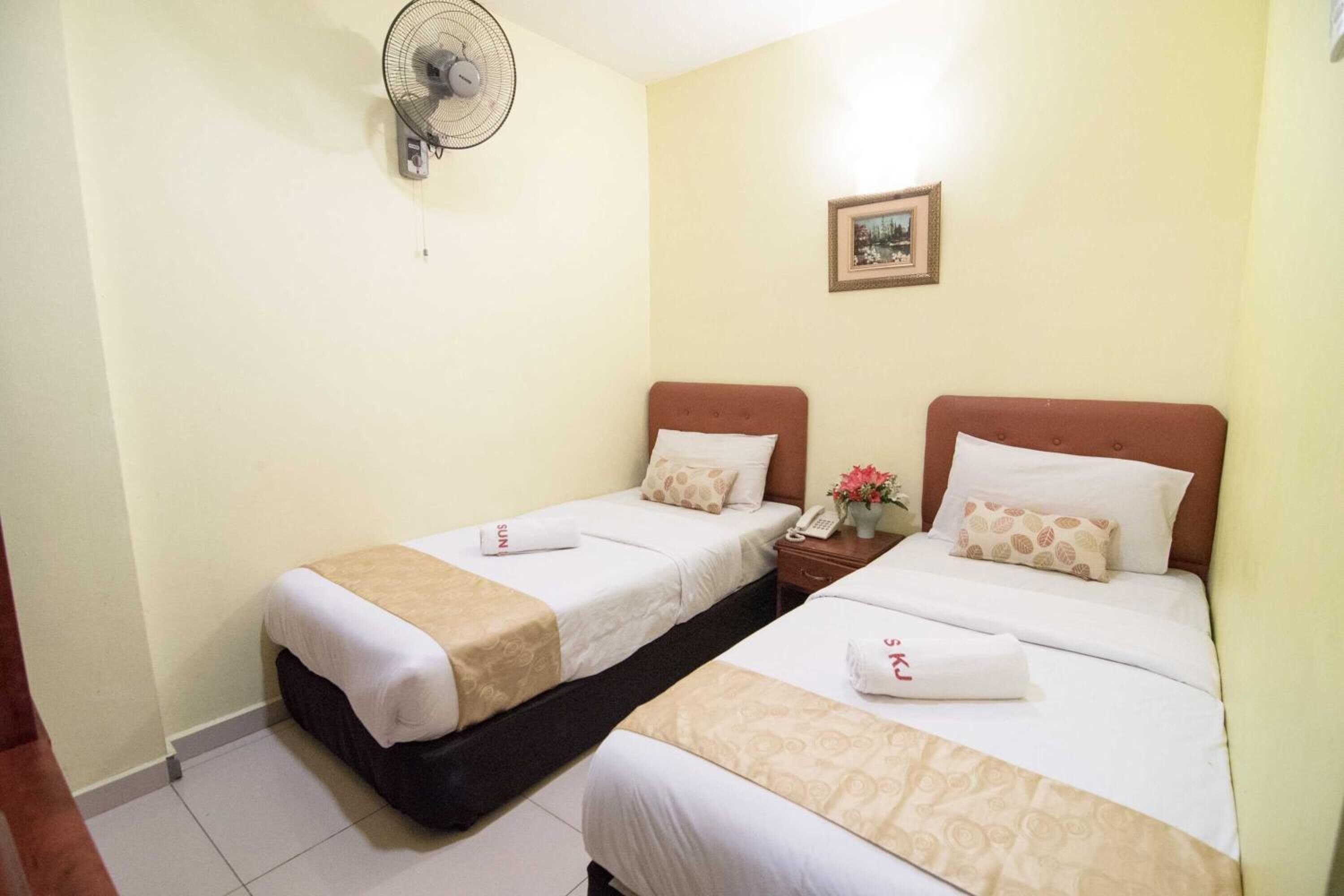 A hotel room with two single beds, each with a white bedspread, a brown throw blanket, and a pillow. There is a small bedside table with a phone and a vase with flowers. A wall-mounted fan is visible, and a framed picture hangs on the wall.