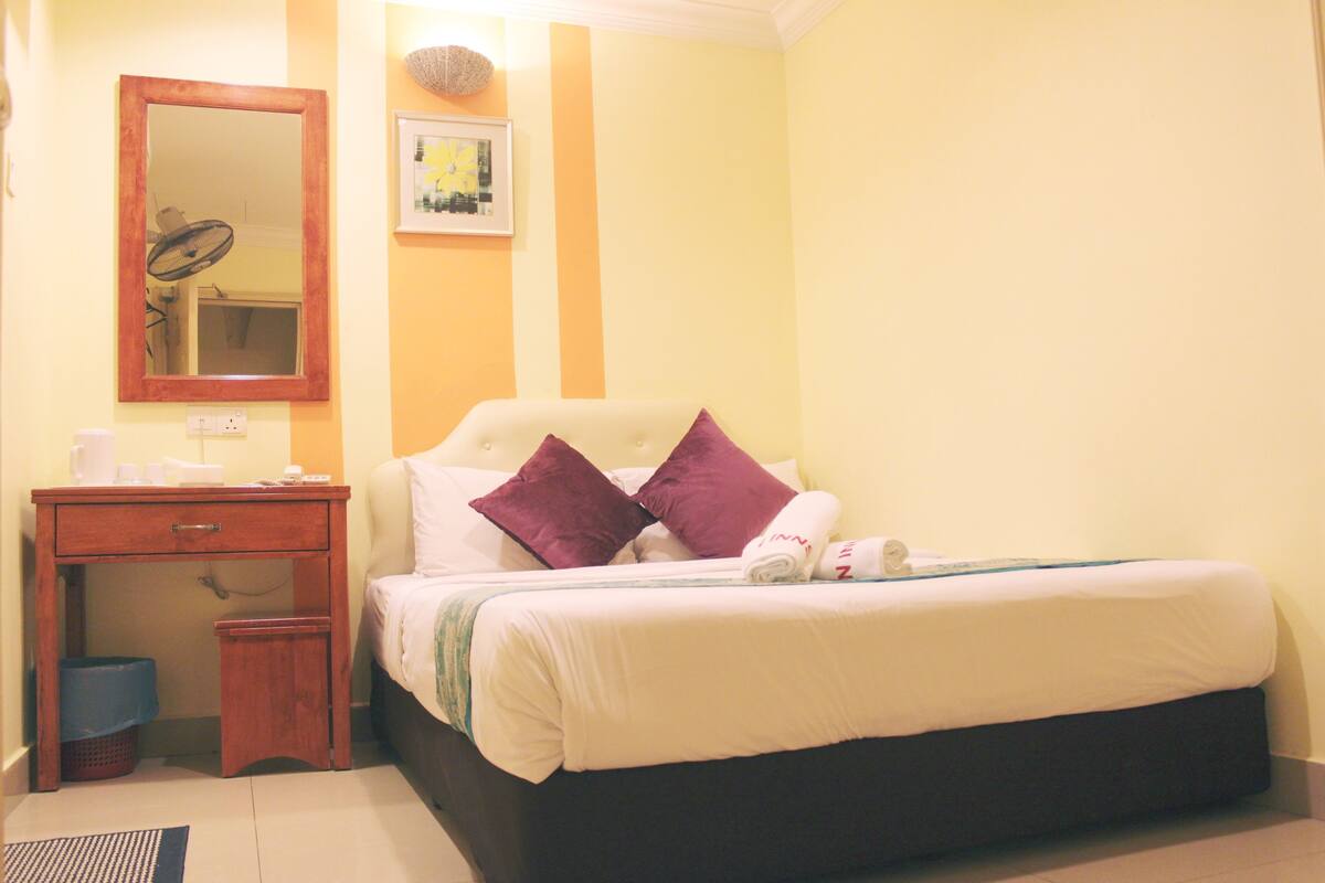 A neatly made bed with white and purple pillows, a wooden nightstand with a mirror, and a small trash bin.