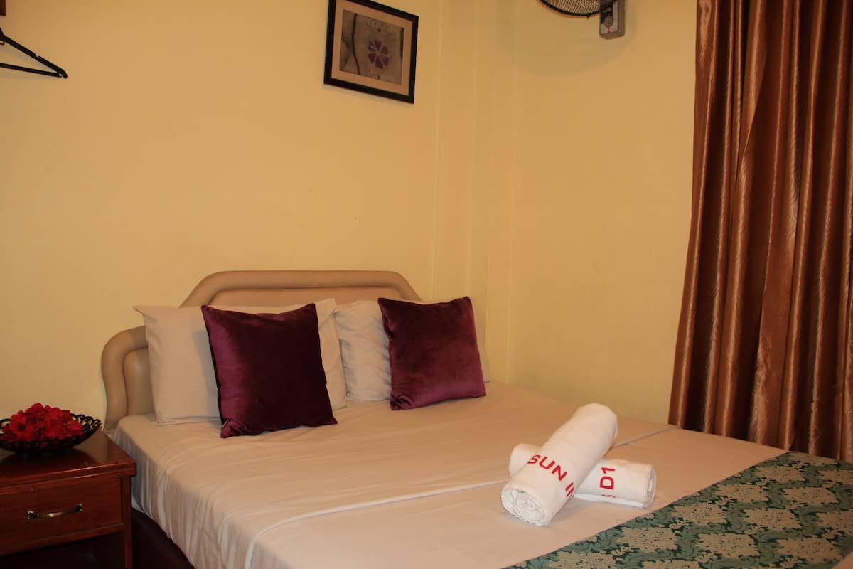 A neatly made bed with a rolled towel, a small bedside table with a bowl of flowers, and a picture on the wall.