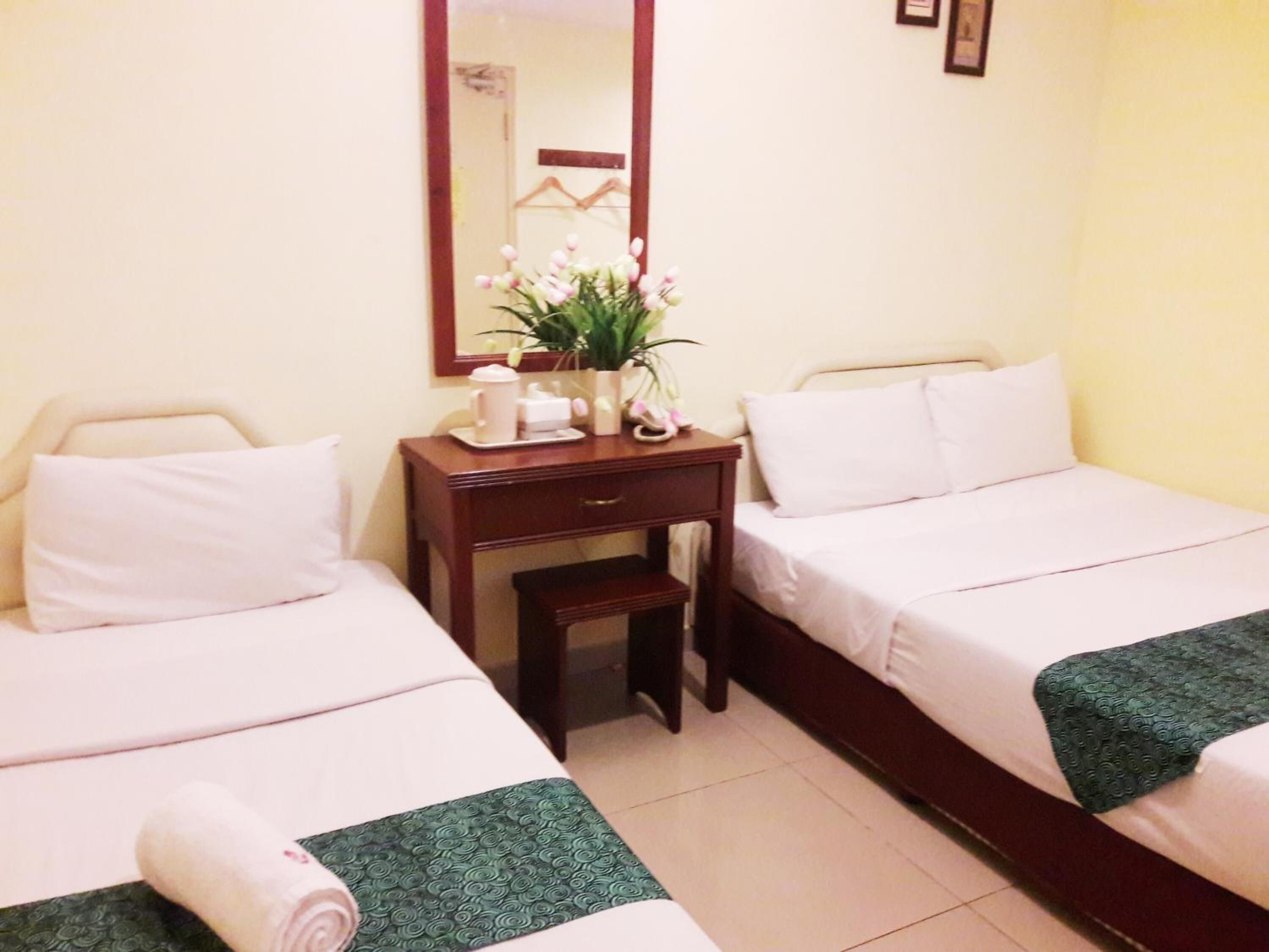 A hotel room with two single beds, a wooden nightstand with a vase of flowers, a mirror, and a small table with a coffee maker.