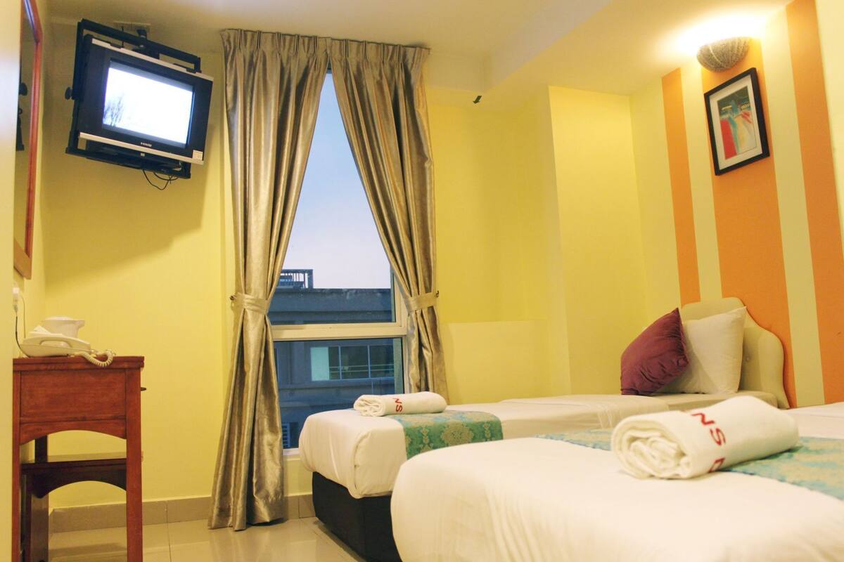 A hotel room with two beds, a TV mounted on the wall, a desk with a telephone, and a window with curtains.