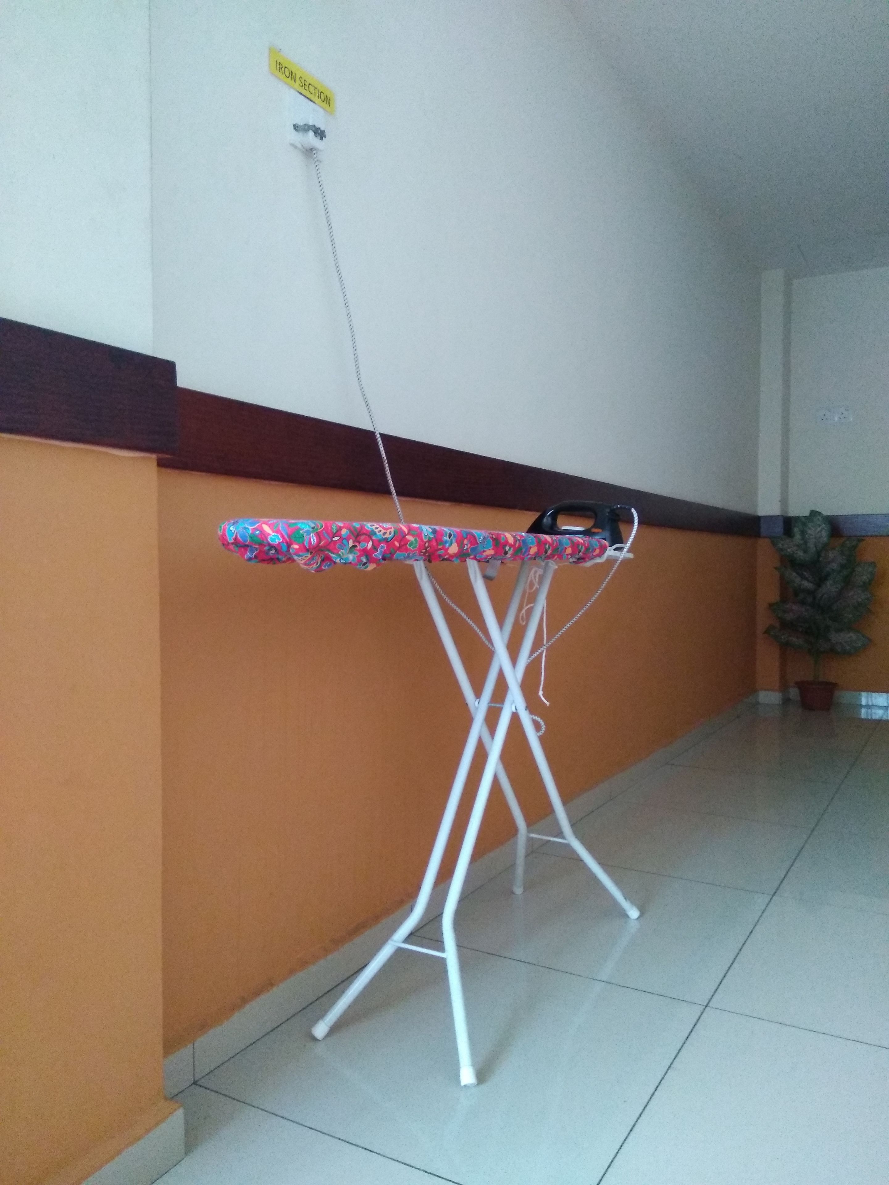 A white ironing board with a colorful cloth cover, placed against a wall with a brown lower section and a white upper section.