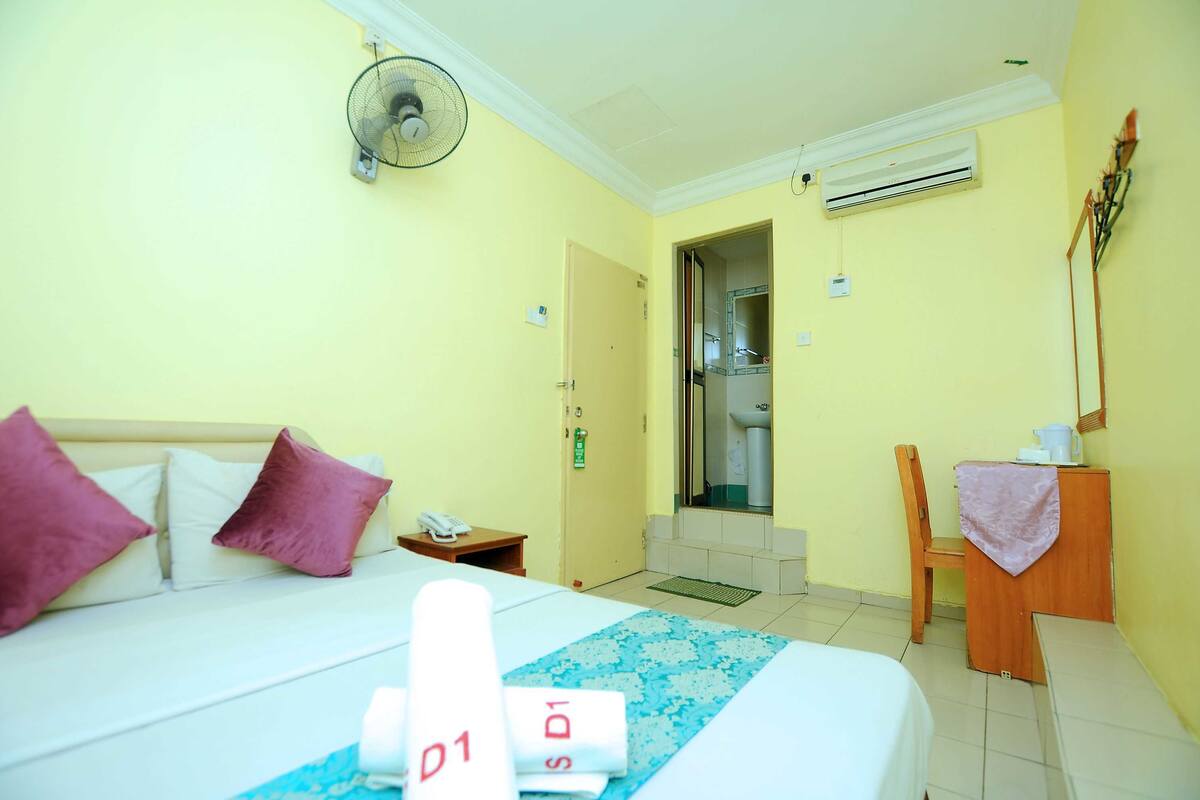 Une chambre d’hôtel équipée d’un lit, d’un bureau, d’une chaise, d’un ventilateur, d’un climatiseur et d’une porte de salle de bain.
