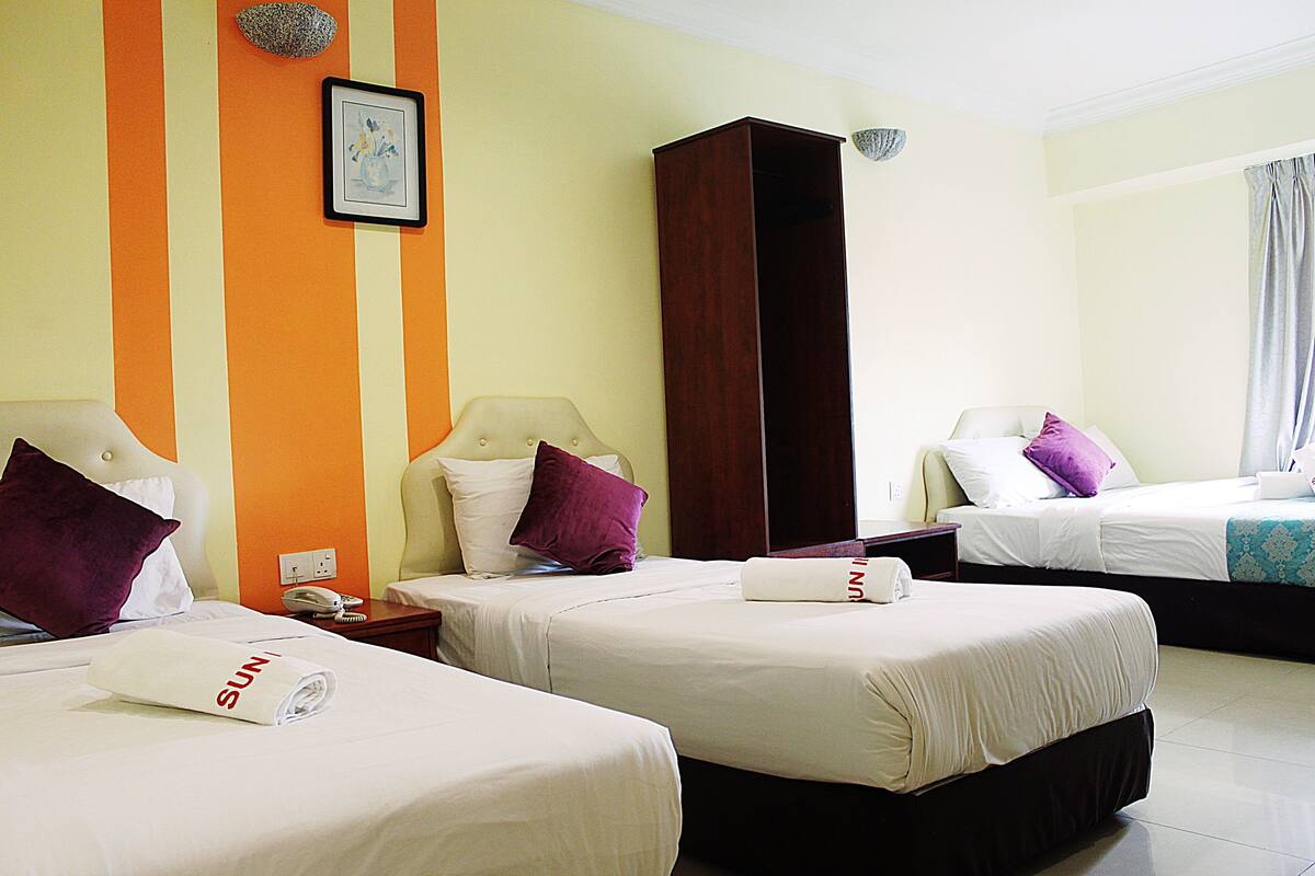 A hotel room with three beds, each with a white bedspread and a rolled towel with "SUN" on it. There is a wooden wardrobe, a nightstand with a phone, and a framed picture on the wall.