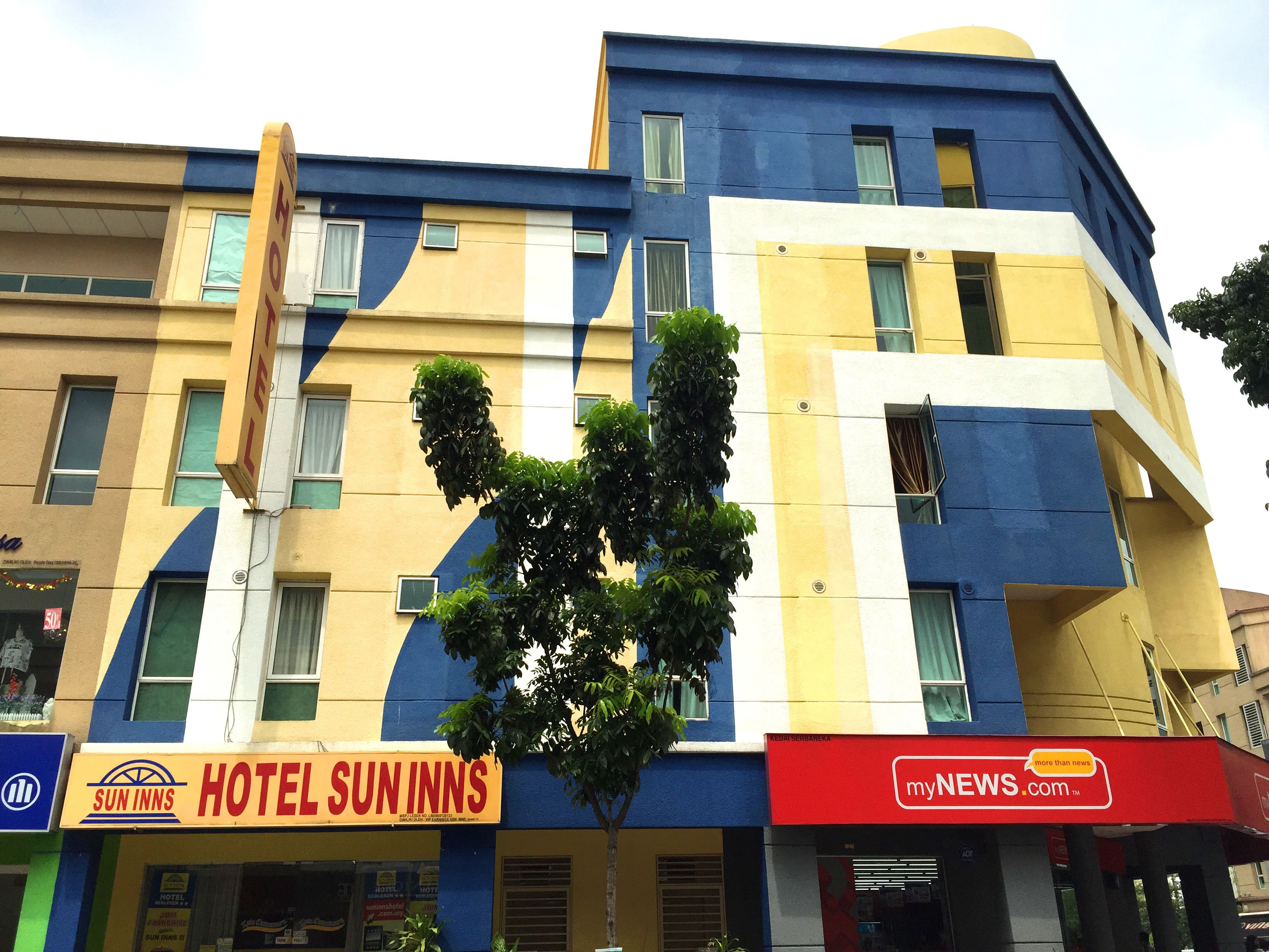 En bygning med flere etager, en gul og blå facade, et synligt skilt med 'Hotel Suninns' og et træ foran.